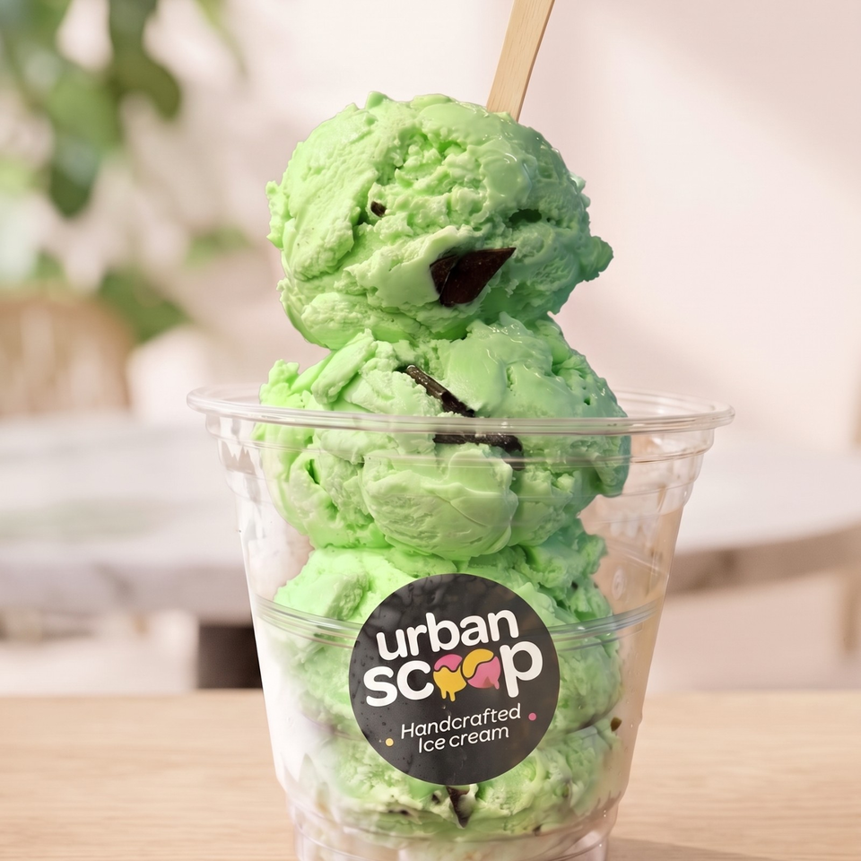 Green mint-chip ice cream in a clear Urban Scoop cup, topped with a scoop and chocolate pieces.
