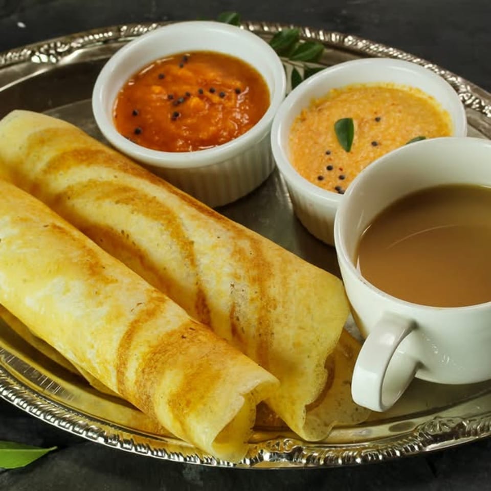 Two golden-brown dosas, two bowls of chutney, and a cup of chai on a silver platter.