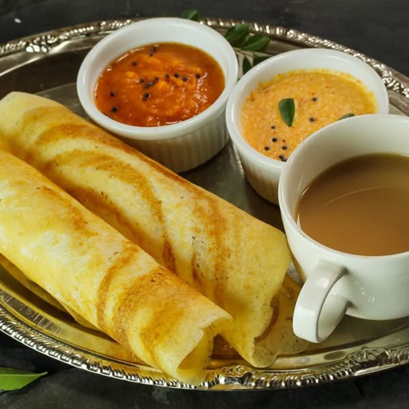 Two golden-brown dosas, two bowls of chutney, and a cup of chai on a silver platter.