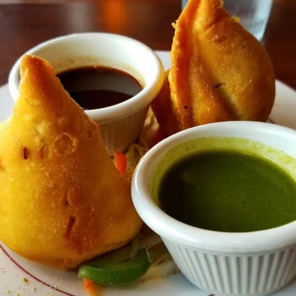 Two golden-fried samosas with small white bowls of dark and green chutneys on a plate.