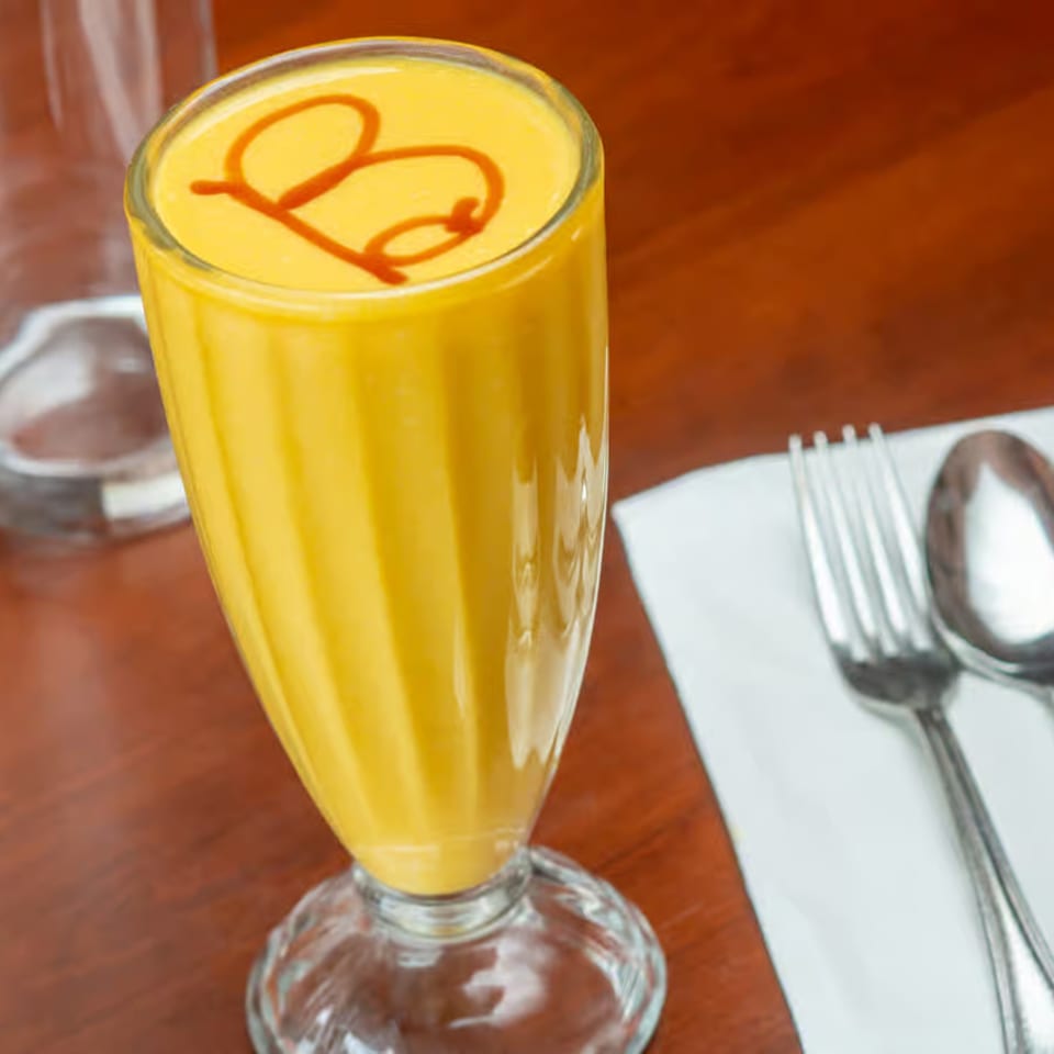 Mango lassi with a decorative "B" swirl on top, next to silverware on a white napkin.