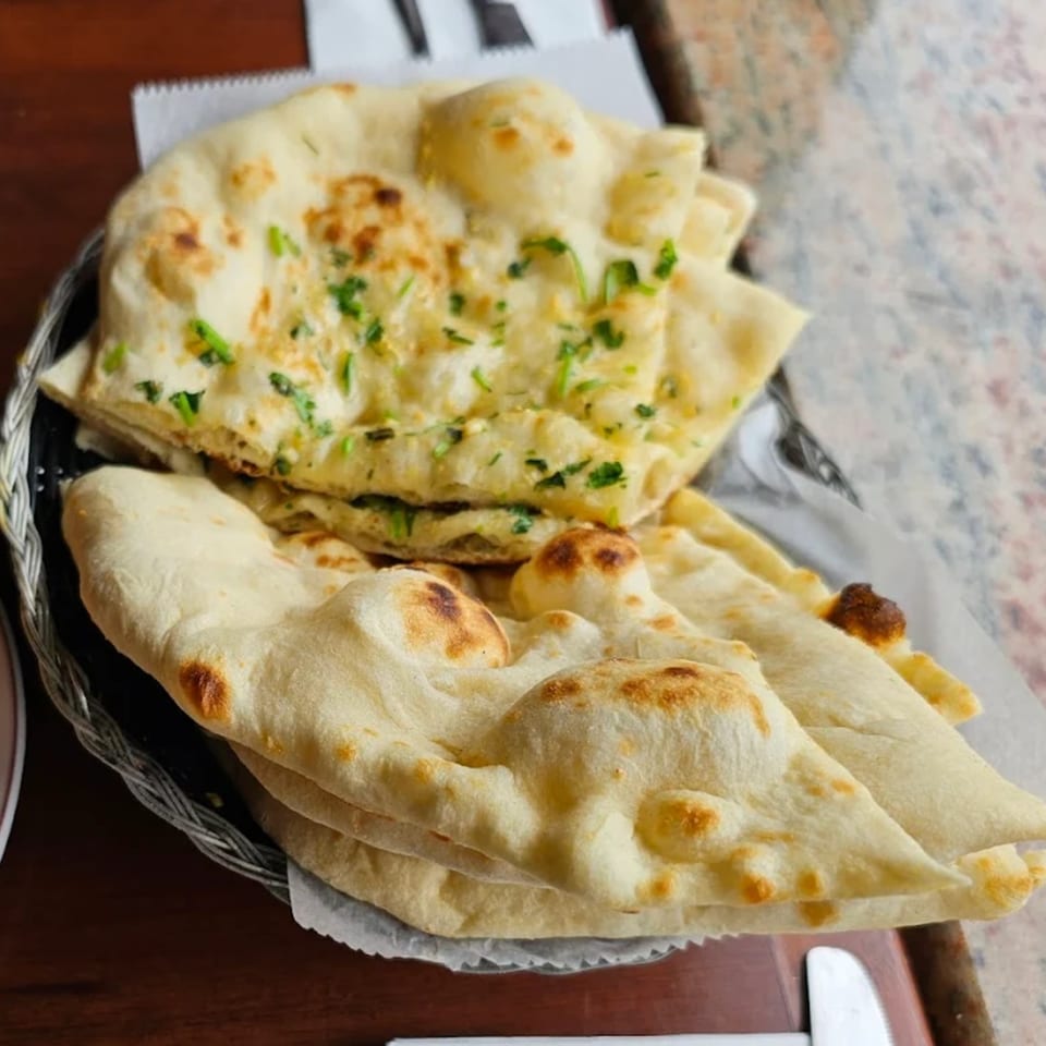 A basket of freshly baked Indian naan bread, some topped with green herbs, on a wooden table.