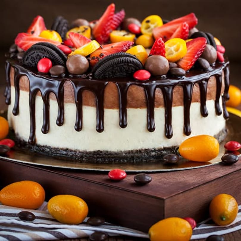 Decadent chocolate drip cake with strawberries, kumquats, cookies, and candies on a wooden surface.